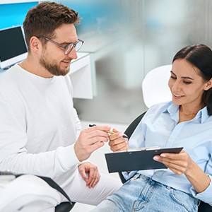 Man in white handing pen to woman holding forms on clipboard in dental chair