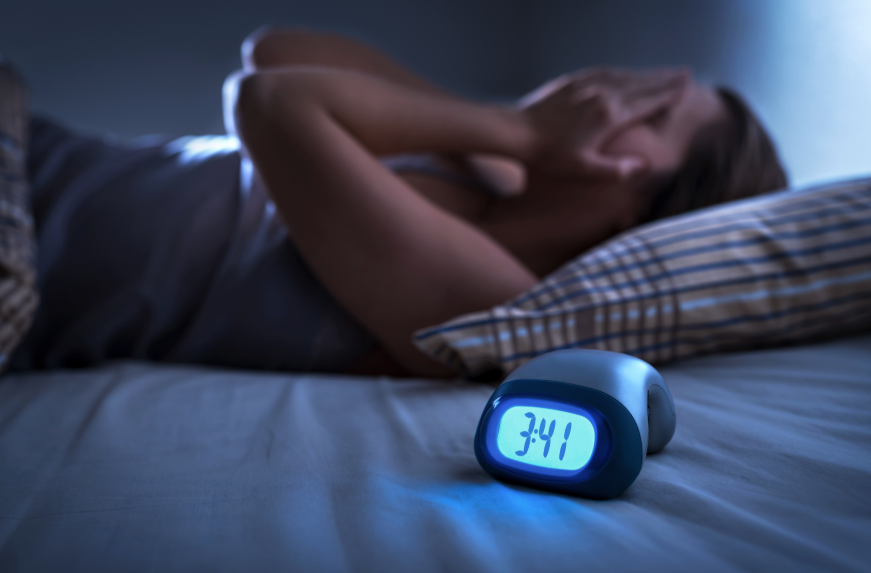 Woman holding face in hands, lying in bed in dark room with alarm clock showing early 3:41 AM